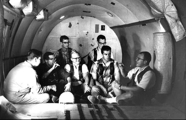 The photo is black-and-white and shows seven white men sitting on the floor of a cylindrical room, smiling and signing to each other. 